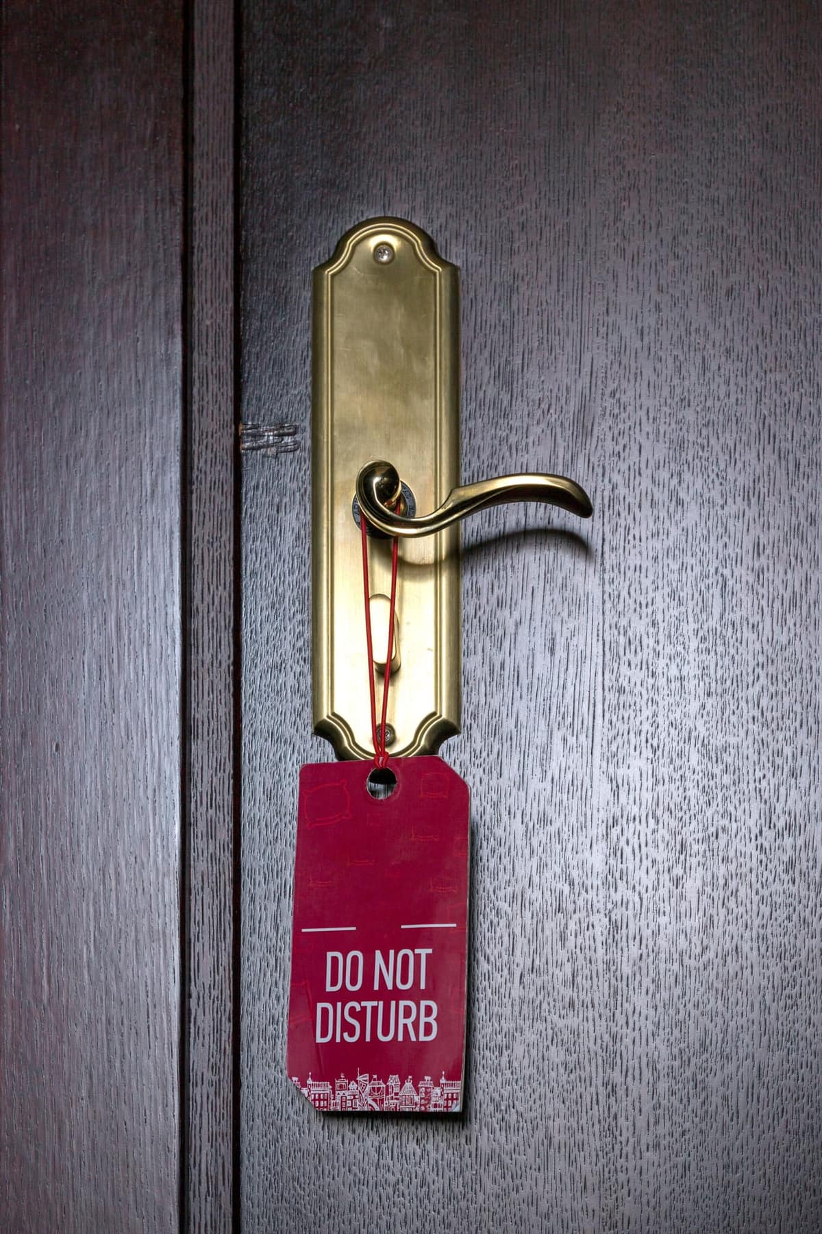 A red DO NOT DISTURB tag hanging from the brass lever handle of a dark wooden door