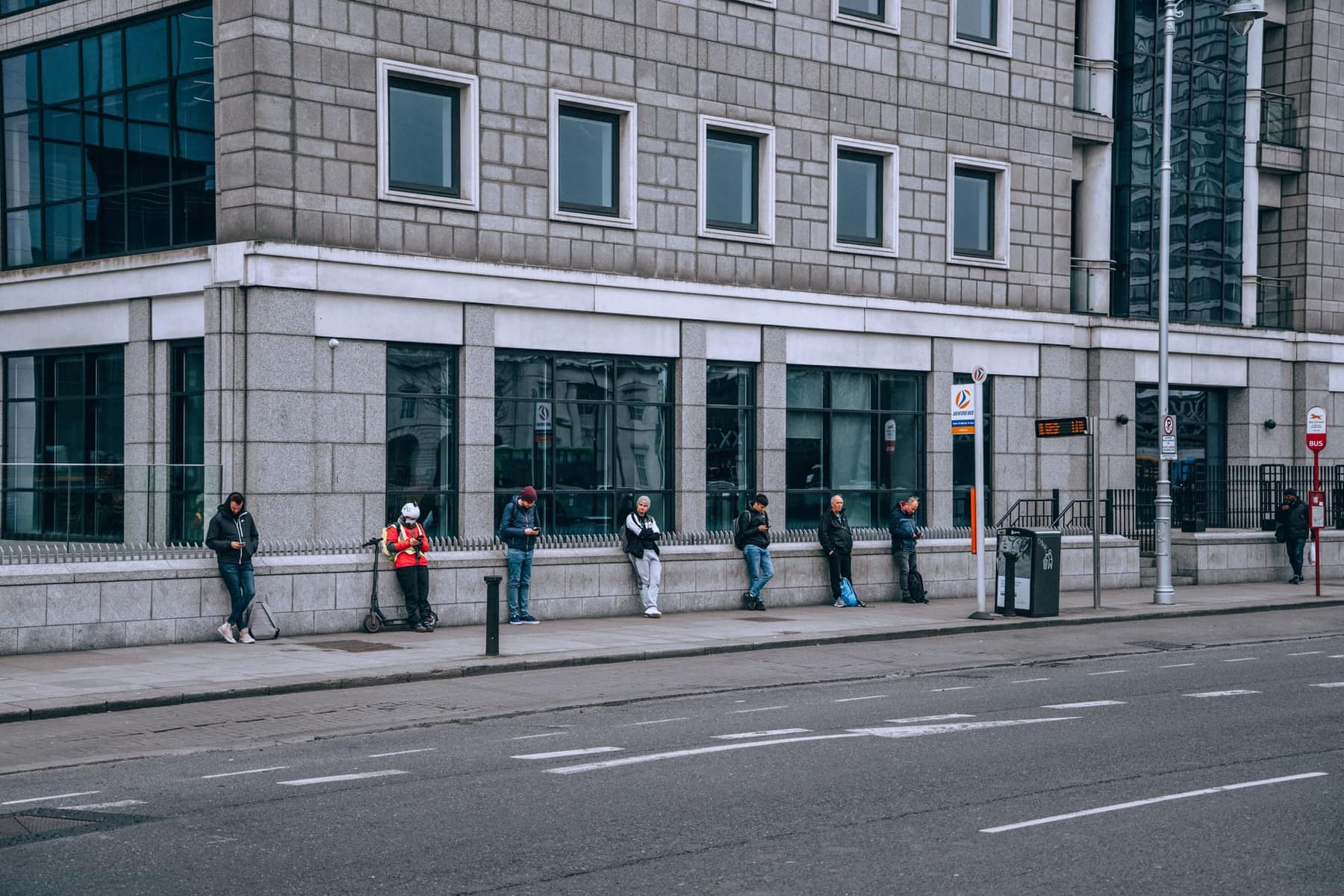 A row of people leaning against a wall at a city bus stop, each waiting alone on their phone