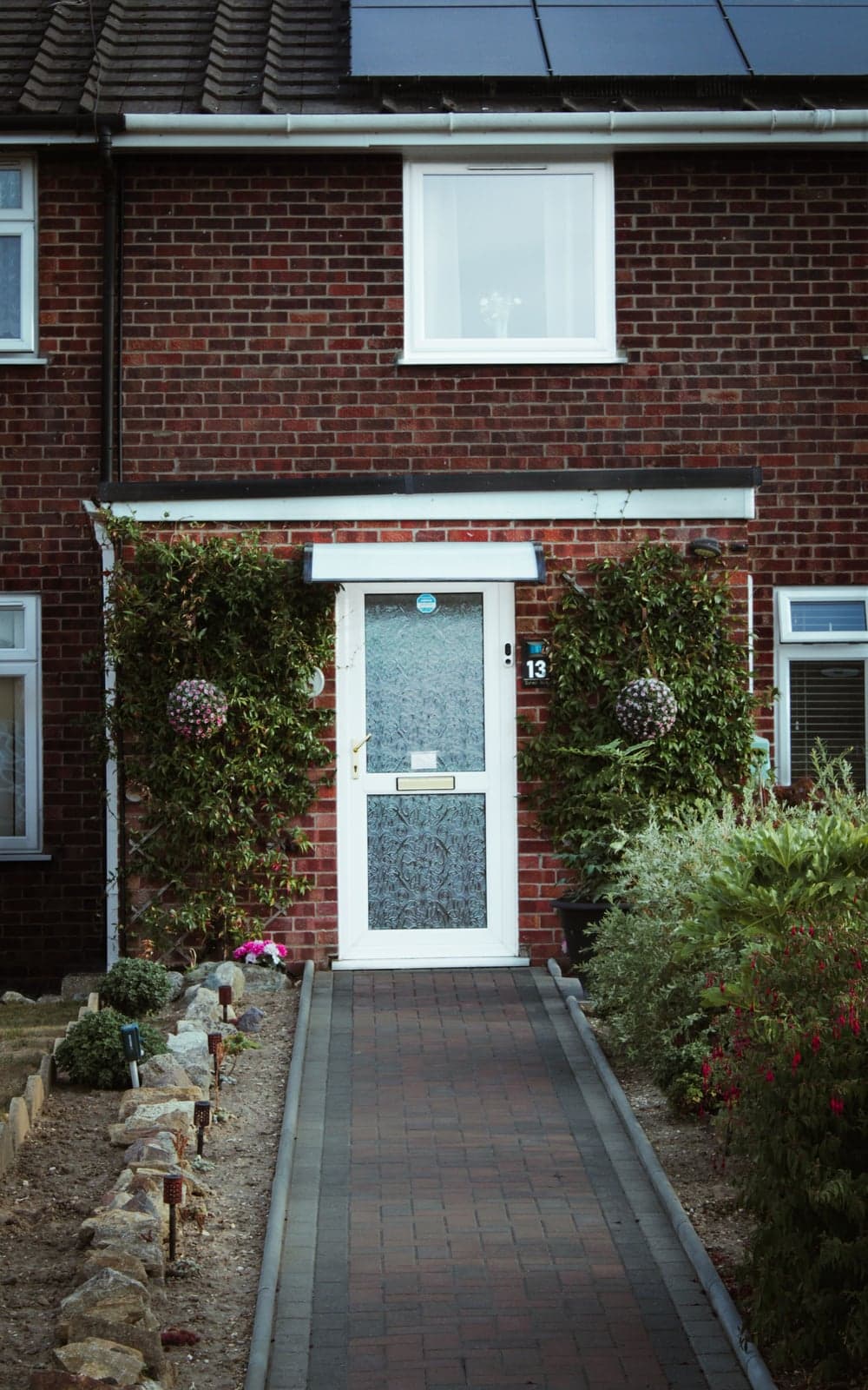 A brick townhouse's white front door numbered 13, flanked by climbing plants and a paved front path